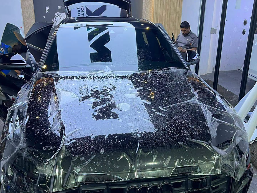 An Audi Q7 bonnet mid-installation with wet paint protection film laid over it, another vehicle and a working figure visible far behind.