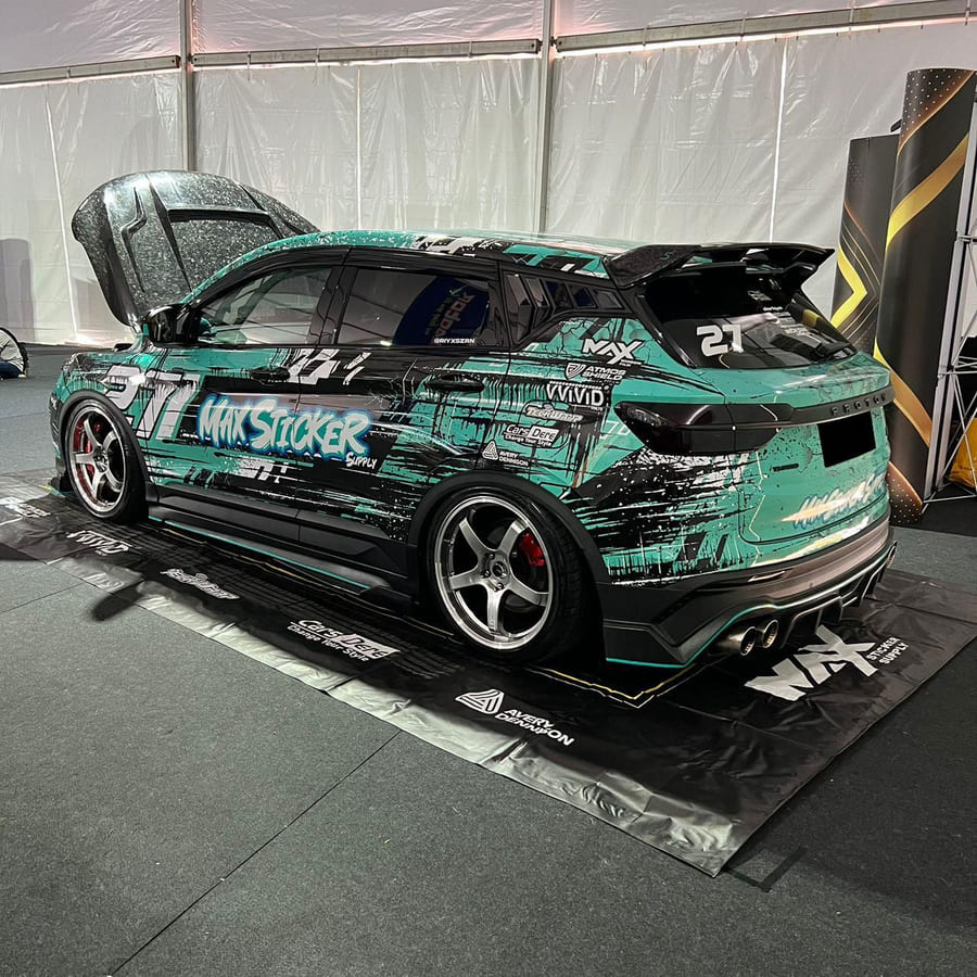 A wide-arch track car in a printed teal-and-black camouflage livery carrying "MAX STICKER" across the rear door and a competition number, inside a plastic-sheeted bay.