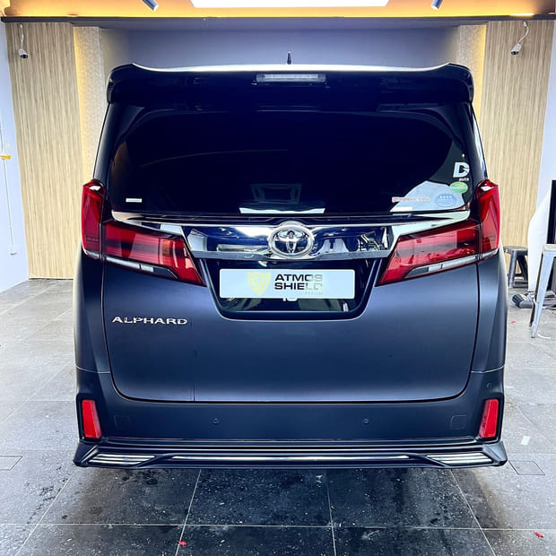 A Toyota Alphard from directly behind in a wood-lined bay, in a dark matte finish.