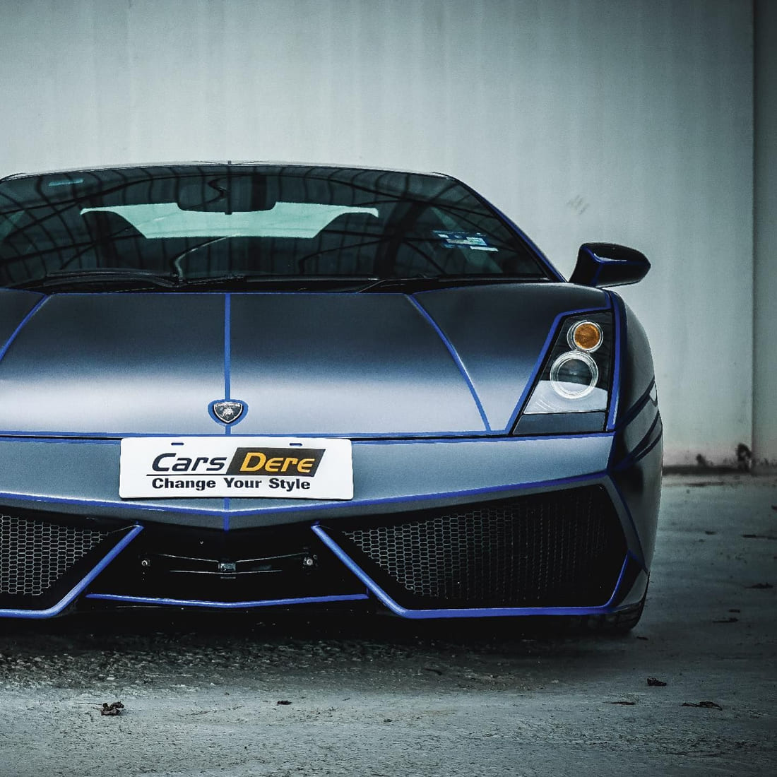 A Lamborghini Gallardo photographed low from the front three-quarter against a plain wall, its body in platinum dark grey with velvet blue accent lines.