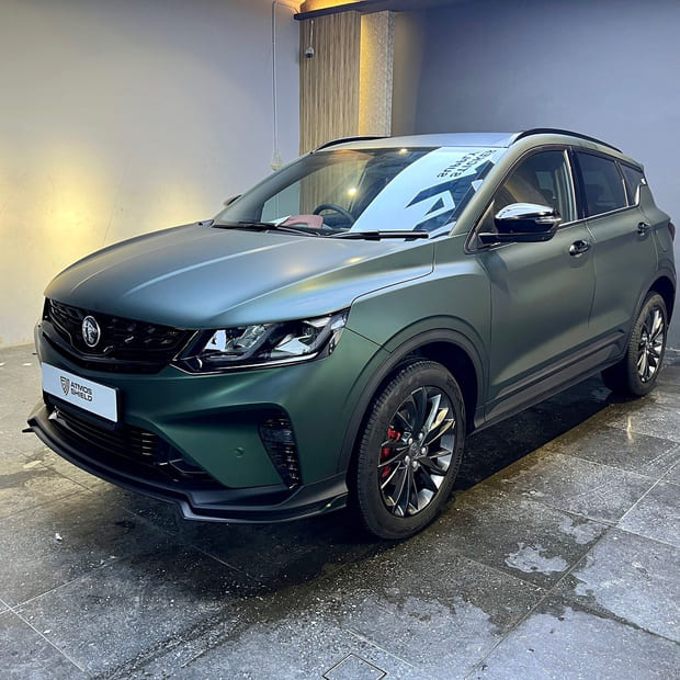 A Proton X50 photographed indoors from the side three-quarter, in a deep metallic green that shifts towards black in shadow.